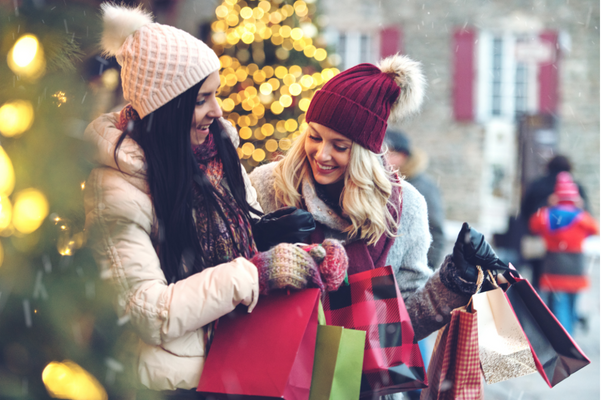 Two women standing outside of a small business in the winter, holding shopping bags and smiling. They are enjoying a day of shopping local.